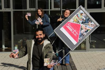 Drei Personen mit Postkarten und PLakaten vor einer Fensterwand